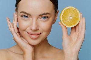 Close up shot of nice looking young woman touches face gently has well cared complexion healthy glowing skin stands bare shoulders holds half of juicy lemon isolated over blue studio background. photo