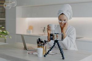 Woman beauty blogger shoots cosmetic vlog looks at camera of smartphone talks with followers applies collagen patches under eyes drinks tea wears white soft bathrobe records video broadcast. photo