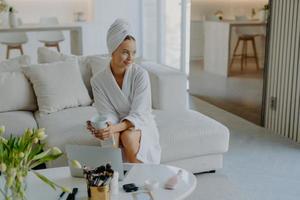 Relaxed thoughtful woman dressed in bathrobe and wrapped towel on head sits on sofa with cup of beverage near table with cosmetic products looks pensive aside poses against cozy home interior photo