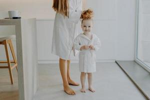 Adorable cute curly girl poses near mother in white soft bathrobe. Unrecognizable mom combs hair of daughter makes pony tail stands bare feet in modern apartment. Hair care and children concept photo