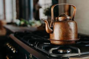 Bronze kettle in modern kitchen. Old vintage teapot on gas stove. Preparing tea. Aluminium teakettle. Sunny daylight from window. photo