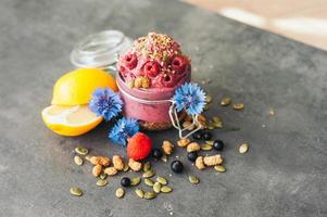 delicioso helado de frambuesa con semillas de calabaza y semillas de cáñamo, decorado con flores azules, grosella negra, limón en rodajas sobre una superficie gris. sabroso postre foto