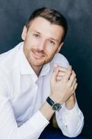 Vertical shot of pleased unshaven male entrepreneur, has bristle, dark hair, keeps hands together, dressed in elegant white shirt, looks at camera with confident expression, isolated over dark wall photo