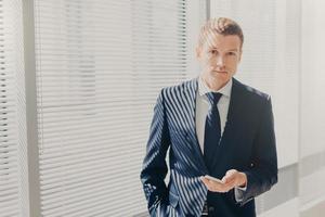 Candid shot of prosperous businessman checks notification on cellular, plans daily schedule, wears formal suit, stands near window, looks directly into camera, has work break. Office routine photo