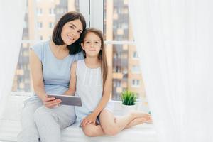 Portrait of beautiful female with dark hair, dressed in casual clothes, holds digital tablet, sits near her daughter, watch something, sit on window sill, play in bedroom. People, lifestyle and family photo