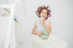 una niña feliz sostiene un peine y un vestido festivo en perchas, tiene rizadores en el cabello, se prepara para la fiesta de cumpleaños con amigos, cuida su belleza y apariencia, aislada de fondo blanco foto