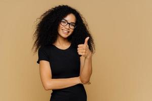 I totally agree. Good looking satisfied Afro American woman keeps thumb up, dressed in casual black t shirt, has crisp hair, wears spectacles, models over brown background. Body language concept photo