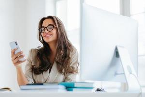 Smiling female in corporate clothes watches funny video via smartphone, sits at desktop in office, uses computer for work, enjoys working process, browses information for project, wears eyeglasses photo