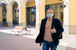 European man wears protective face mask while coronavirus and pandemic, carries rucksack, poses outdoor at bus station, travels during quarantine. Covid-19 virus, epidemic outbreak, public place photo