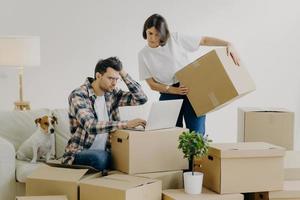 Moving concept. Frustrated young man uses modern laptop computer for searching necessary information online, woman replaces big carton boxes with personal belongings. New home and life concept photo