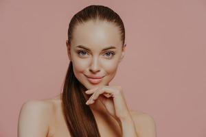 Skin care and anti aging procedures. Confident brunette young woman keeps hand under chin, has combed hair, clean fresh skin, enjoys beauty treatments, poses with naked body over pink background photo