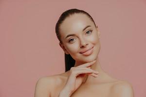 Charming beautiful woman with pony tail, touches perfect soft skin after cosmetology procedures, using beauty cream, stands with bare shouldes, poses against pink background. Natural as she is photo