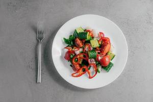 Top view of fresh vegetable salad prepared of red pepper, radish, tomatoes, cucumbers and parsley in white bowl, fork near. Vegetarian dish concept. Healthy nutritious spring salad photo