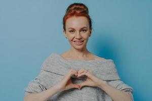Kind smiling red haired woman showing heart gesture on chest isolated on blue wall background photo