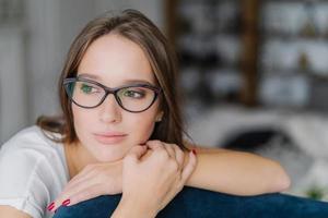 Thoughtful dark haired European female in spectacles looks aside, leans at back of couch, contemplates about future, feels lonely, enjoys domestic atmosphere, poses against blurred background photo