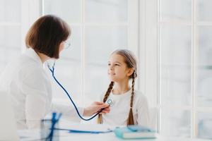 Lovely small girl listens consult of professional experienced doctor who listens her lungs with stethoscope, comes on medical checkup appointment. Children healthcare and clinic visit concept photo
