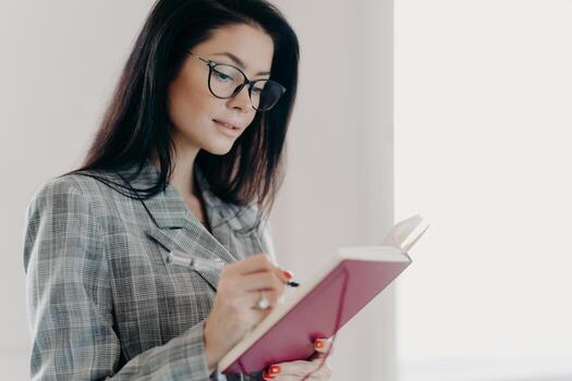Sideways shot of hard working executive woman in spectacles, formal checkered suit, writes down information in notepad, makes list to do in diary, notes point goals, poses indoor, white background photo