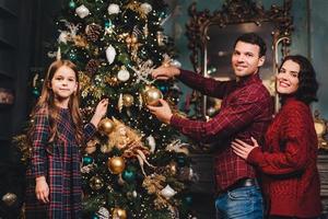 familia amistosa de padre, madre e hija decora el árbol de año nuevo, contentos de tener próximas vacaciones de invierno, disfrutar del ambiente doméstico. familia de tres miembros ocupada con decoraciones foto