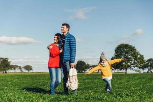 Young couple in love stand on green grass, embrace and look pensively. Playful small child enjoys freedom, runs on field, plays near her parents, has good mood and recreation. Peope and nature photo