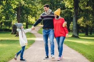 Outdoor portrait of affectionate family walk in park, wear warm knitted clothes. Handsome young man holds daughters hand, looks with happy expression at her. Rest and relaxation concept photo