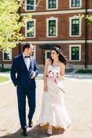 Beautiful young bride in white wedding dress, holds bouquet, has talk with her future husband, have walk together, go to registry office. Lovely married couple rejoice special day in their life. photo