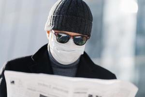 foto de un hombre atento que usa gafas de sol negras, sombrero y máscara médica en la cara durante la cuarentena y el brote de coronavirus, lee la prensa, descubre noticias del periódico, se encuentra en un fondo borroso