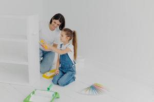 Little girl learns how to paint with roller, stands on knees, her mother tells how to do it correctly, pose in spacious white room, choose color from palette samples. Home makeover, decoration concept photo