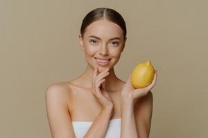 Cosmetology skin care and beauty concept. Pleased brunette young woman wrapped in bath towel holds lemon for making facial mask has refreshed clean skin after taking shower and spa procedures photo