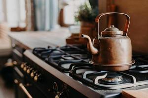 Old aluminium kettle boilng on gas stove in kitchen against cozy blurred background. Antique item made of copper metal. Vintage style photo