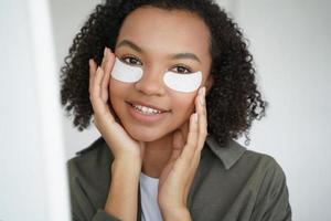 Anti wrinkle procedure. Happy afro girl is applying eye patches. Skincare of hispanic young woman. photo