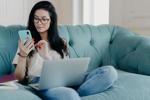 Serious brunette female freelancer works remotely on laptop computer, sends text messages via smartphone connected to wireless free internet sits crossed legs on comfortable sofa surfs social networks photo