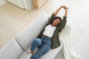 tranquila joven afroamericana acostada, estirando el cuerpo, relajándose en el sofá en la sala de estar en casa foto