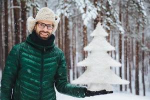 el hombre positivo sonriente usa anteojos, sombrero y anorak, sostiene un abeto artificial, se para contra los árboles cubiertos de nieve, mira directamente a la cámara con expresión feliz, pasa tiempo en el clima helado foto