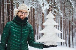 Smiling positive male wears glasses, hat and anorak, holds artificial fir tree, stands against trees covered with snow, looks directly into camera with happy expression, spends time on frosty weather photo