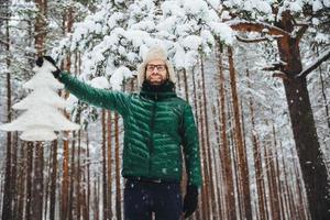 toma horizontal de un atractivo hombre barbudo que se alza contra los árboles de invierno en el bosque, sostiene un abeto artificial blanco, expresa un estado de ánimo festivo y se prepara para las vacaciones de invierno. gente, clima, temporada foto