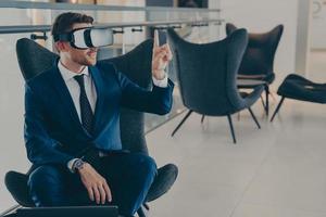 Young CEO sitting in office centre lobby with VR headset goggles, pointing with forefinger up in air photo