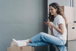 Happy attractive hispanic woman is sitting on the sill with boxes and texting on mobile phone. photo