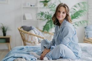 Indoor shot of pretty dark haired woman sits crossed legs on bed, dressed in nighclothes, white socks, looks direcltly at camera, feels relaxed while listens calm music in earphones. Rest and day off photo