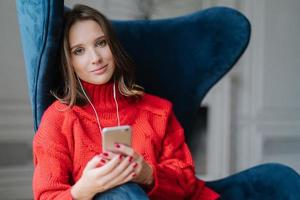 Photo of charming female sits in comfortable armchair, dressed in sweater, listens audio book, connected to earphones, uses modern cell phone and high speed internet connection. Leisure concept
