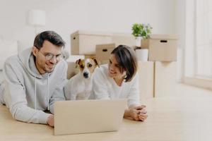 gente y nuevo concepto de apartamento. el feliz esposo y la esposa se relajan en el piso, usan una computadora portátil moderna para navegar por la red, posan en su nuevo hogar o apartamento, su perro posa cerca, descansan poco foto