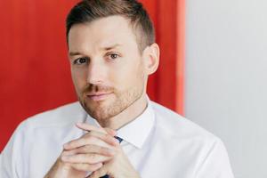 Cropped image of confident young male employee with bristle, keeps hands pressed together, dressed in elegant white shirt, contemplates about opening new firm, isoated over red and white wall photo