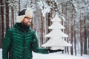 un hombre sonriente de mediana edad mira con expresión feliz a un pequeño abeto blanco artificial, va a celebrar las vacaciones de año nuevo, pasa la mañana al aire libre en el bosque durante el clima nevado foto