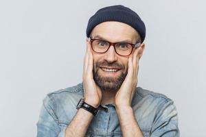 Portrait of attractive young man with thick beard and mustache looks with delighted expression, has white perfect teeth, poses in studio against blank copy space. People and positive emotions photo