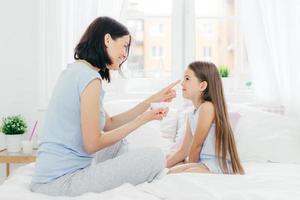 Affectionate mother applies cream on daughters nose, sits together at bed, takes cake of healthy skin and good appearance, have beauty procedure at home. Family, skin care and beauty concept photo