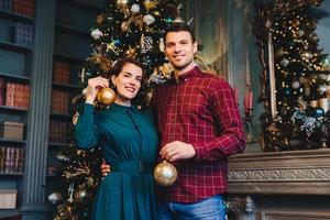 Handsome man with stubble embraces his beautiful wife, hold glass balls in hands, decorate New Year tree together, prepare for holidays. Smiling young beautiful female and her husband at home photo