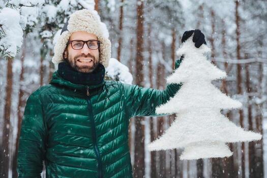 foto al aire libre de sonrientes orejas masculinas de mediana edad sin afeitar gorra cálida y anorak verde, sostiene un abeto artificial, disfruta de hermosos paisajes durante las vacaciones de invierno, tiene una mirada optimista. clima nevado