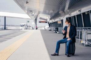 European man sits at suitcase on bus platform station, tries to find transport schedule online on smartphone, waits for transport, travels during dangerous pandemic situation, wears medical mask