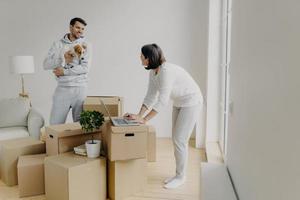 Busy woman tries to find information in laptop computer, buys furniture online, man stands with dog on hands, smiles and talks to wife, surrounded with pile of cardboard boxes have to unpack property photo