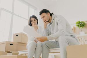 Upset couple stressed by bad news, receive high taxes, looks attentively at papers, read paper letter, have to pay much for flat, move in other apartment, pack belongings in cardboard containers photo