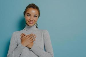 Attractive young smiling woman holding folded hands near heart on chest while posing in studio photo